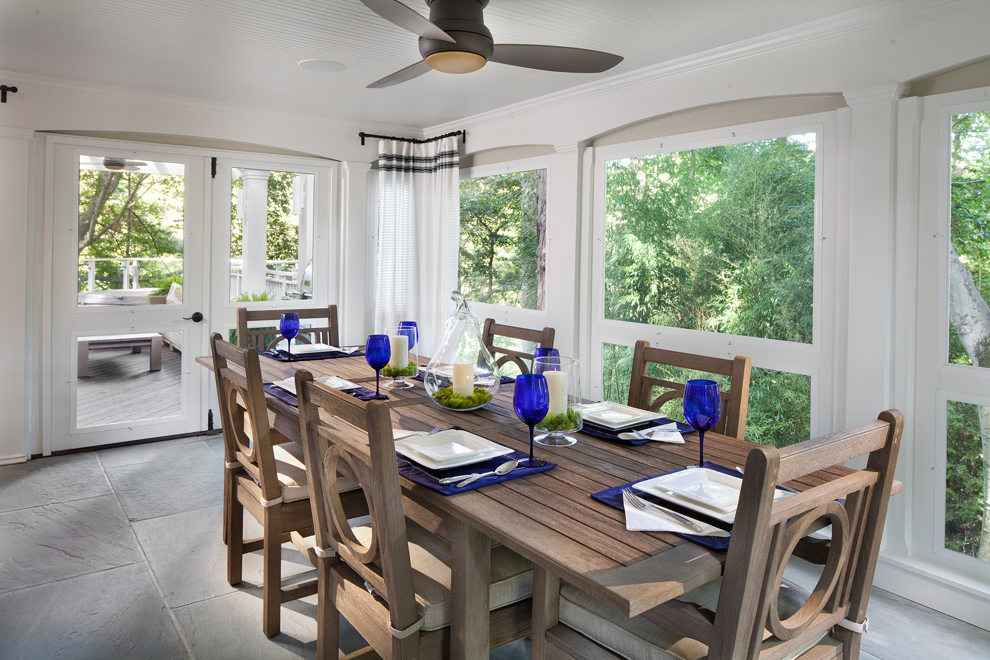 Screened-in porch with ceiling fan, wood dining table, chairs and a blue pop of color place settings