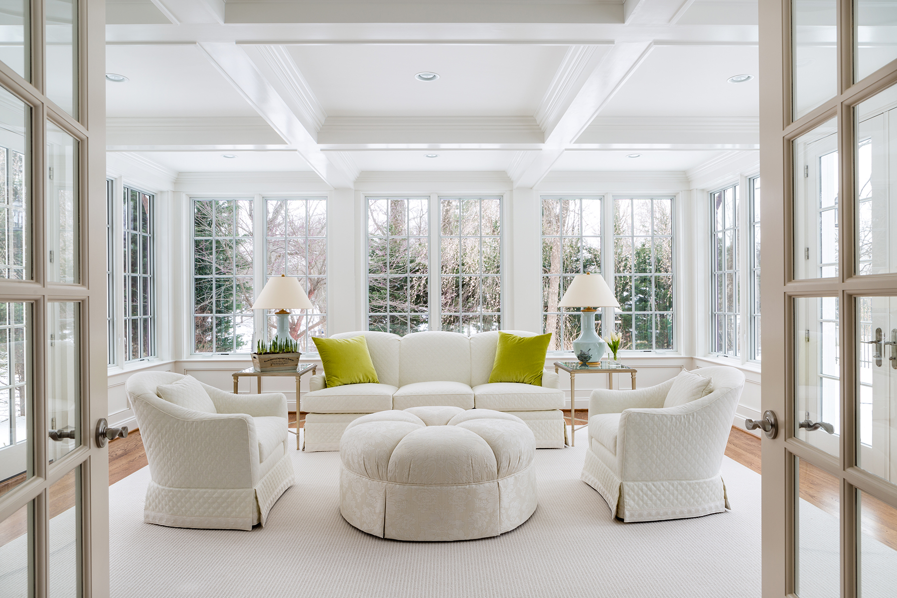 Interior designed light colored living room with white upholstered sofa and accent chairs. Pop of color green pillows to enhance the greenery outside the windows