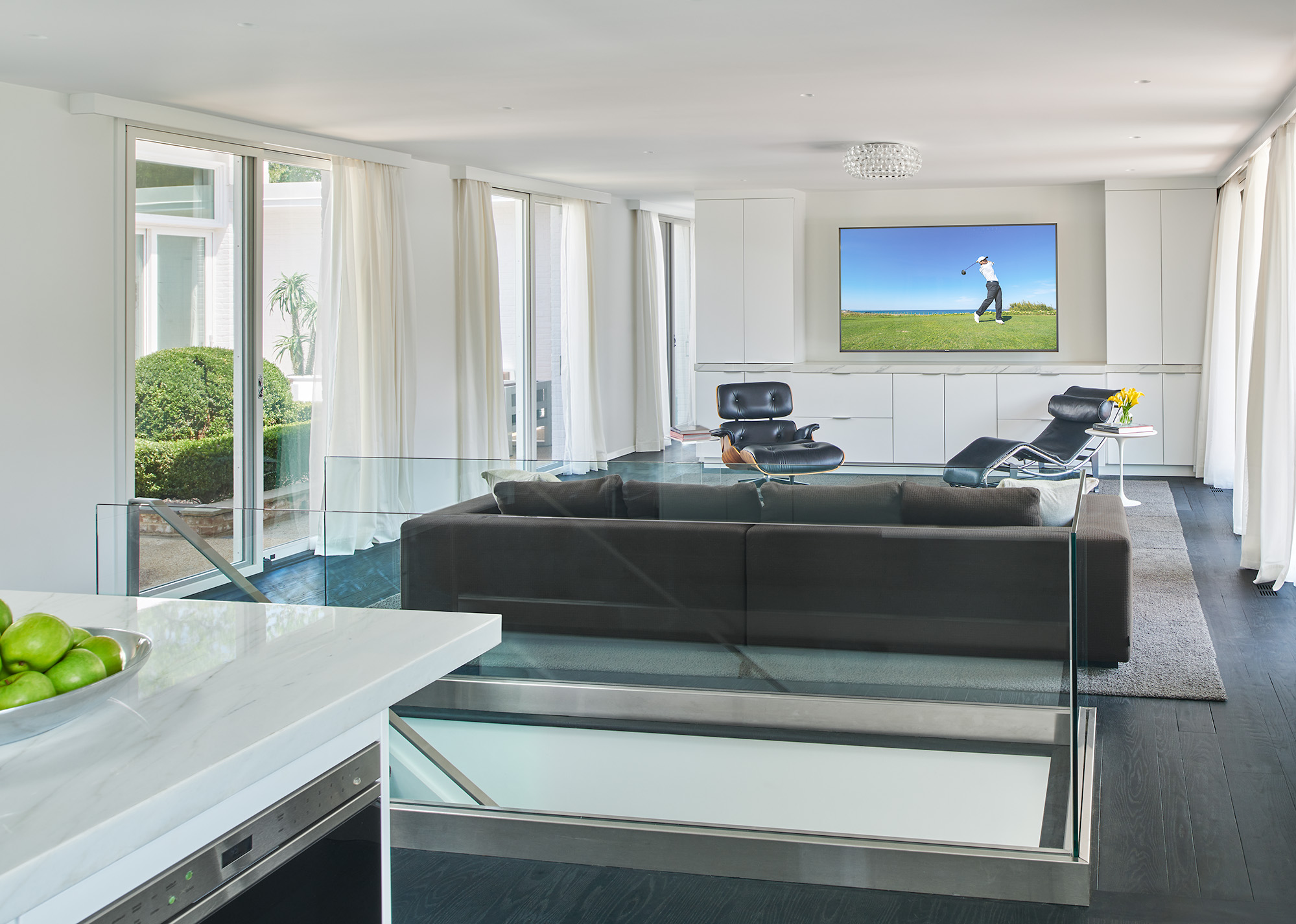 Modern living room with long dark couch, black accent chairs atop a gray area rug. Built in white cabinetry surrounding a large tv